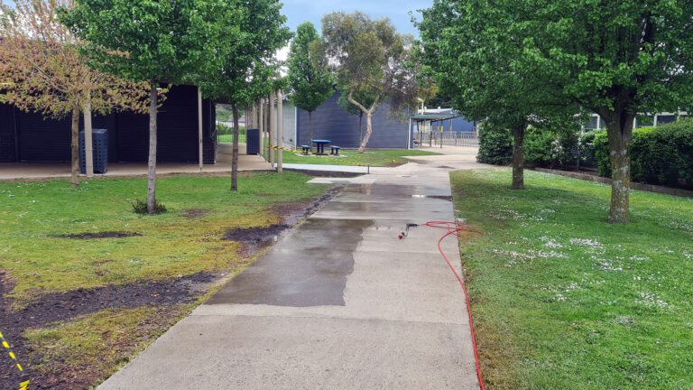 Concrete footpath in the process of being pressure cleaned by AAA Brick Cleaning. A small patch in center is still dirty. Surrounds are green grass, trees and a school building.